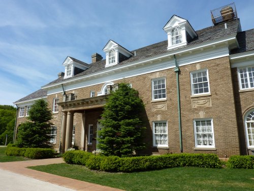 A large brick building with a grand entrance, featuring white windows and manicured greenery in front.