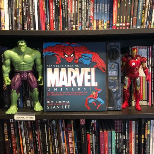 A shelf filled with DVDs and graphic novels, featuring Hulk and Iron Man figures beside a book titled "Stan Lee's Amazing Marvel Universe."