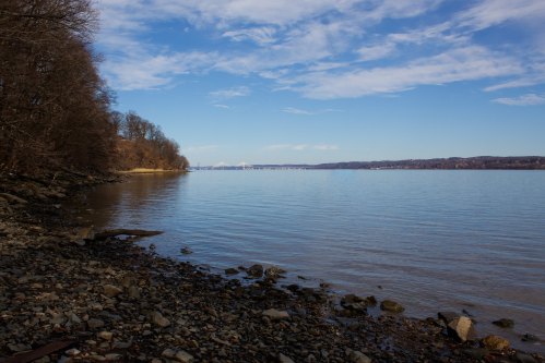 A serene river scene with rocky shorelines, trees, and a clear blue sky reflecting on the calm water.