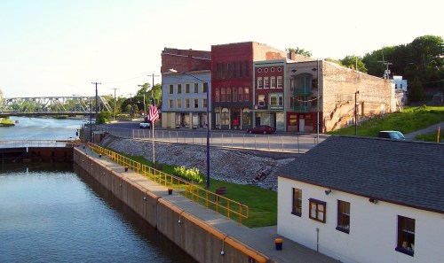 A riverside view of historic buildings and a small dock, with an American flag and a bridge in the background.