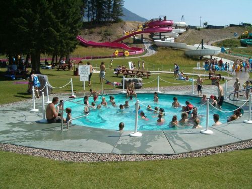 A crowded water park scene with children and adults in a pool, surrounded by slides and grassy areas.
