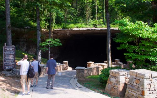 Visitors stand on a path near the entrance of a large cave surrounded by lush greenery and stone walls.