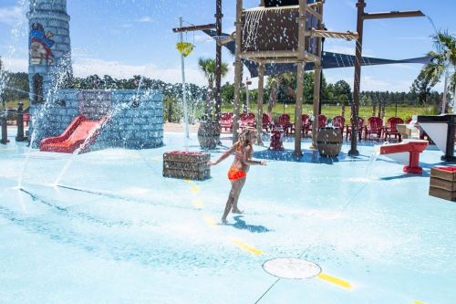 A child in an orange swimsuit plays in a splash pad, surrounded by water features and colorful seating.