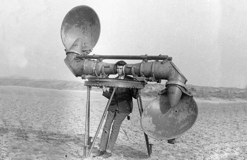 A man stands beside a large, vintage sound device with two horn-like speakers in a barren landscape.