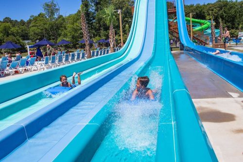 Two children slide down bright blue water slides, splashing into the pool below on a sunny day at a water park.