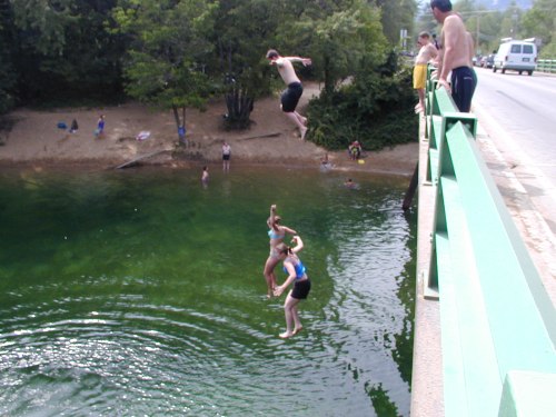 A group of people jumping off a bridge into a clear body of water, with others swimming and relaxing nearby.