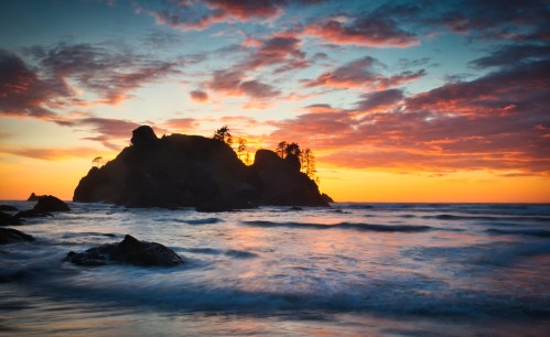 A serene beach scene at sunset, featuring rocky shores and silhouetted trees against a colorful sky.