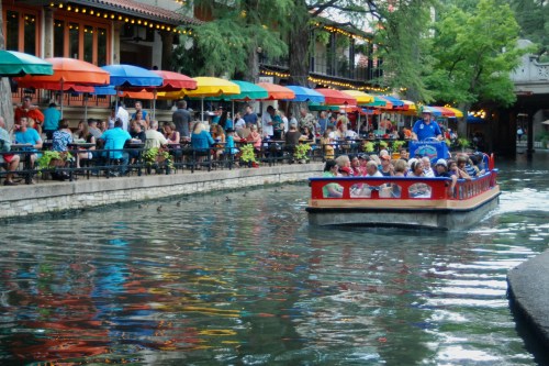 A colorful boat glides through a lively canal lined with diners under vibrant umbrellas.
