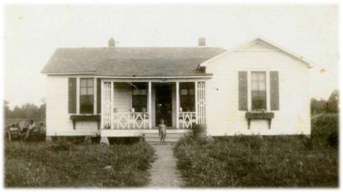A vintage photo of a white house with a porch, surrounded by grass, and a child standing in front.