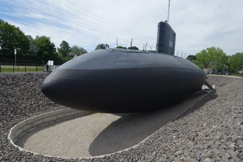 A black submarine displayed on a gravel bed, surrounded by greenery and power lines in the background.