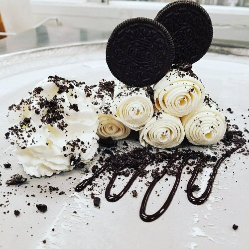 A dessert featuring rolled white chocolate, whipped cream, Oreo cookies, and chocolate drizzle on a silver plate.
