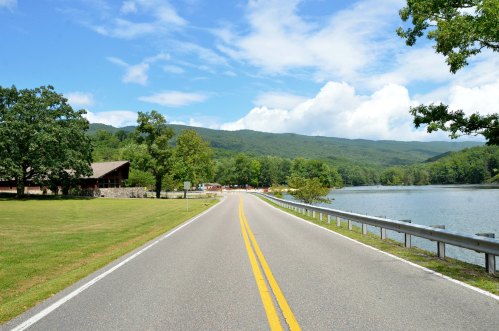 A scenic road lined with trees, leading to a lake with mountains in the background under a blue sky with clouds.