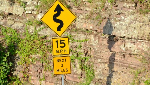 Winding road sign with a speed limit of 15 MPH for the next 3 miles, set against a rocky background.
