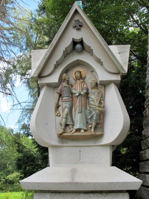 A stone monument featuring a relief of three figures, surrounded by greenery and a clear blue sky.