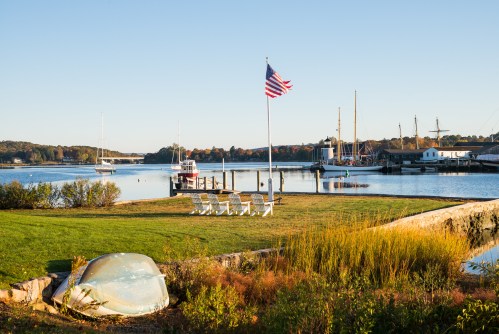 A peaceful waterfront scene with a flag, chairs, a boat, and sailboats in the background under clear blue skies.