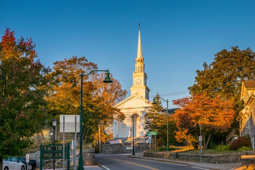 A white church with a tall steeple stands at the end of a tree-lined street, surrounded by autumn foliage.