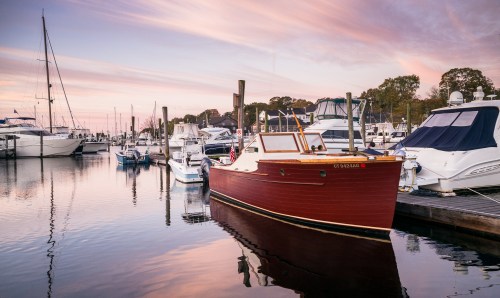 A red boat docked at a marina during sunset, surrounded by white yachts and calm water reflecting the sky.