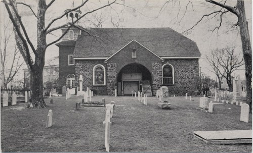 Historic stone church with a graveyard, featuring tombstones and large trees, set in a serene landscape.