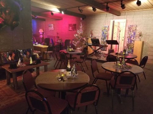 Cozy venue with tables set for an event, decorated with a Christmas tree and soft lighting, featuring a stage and musical setup.