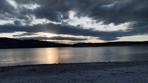 A serene lake at sunset, with dark clouds and a reflective water surface, framed by distant hills.