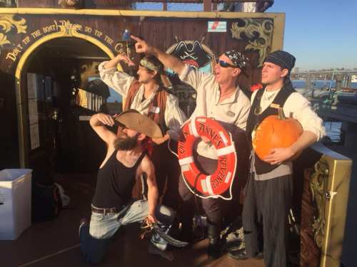 Four pirates pose on a ship deck, one holding a pumpkin, with a life preserver and playful expressions.