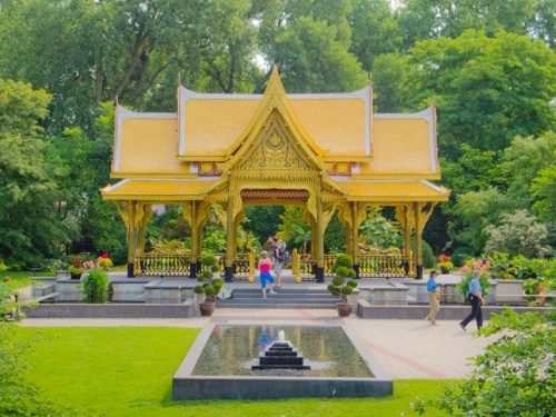 A golden pavilion surrounded by lush greenery and a reflecting pool, with visitors exploring the serene garden.