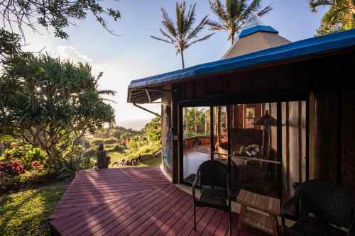 A cozy cabin with a blue roof, surrounded by lush greenery and palm trees, overlooking a scenic landscape.