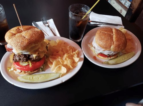 Two large burgers with toppings on plates, accompanied by chips and pickles, with drinks in the background.