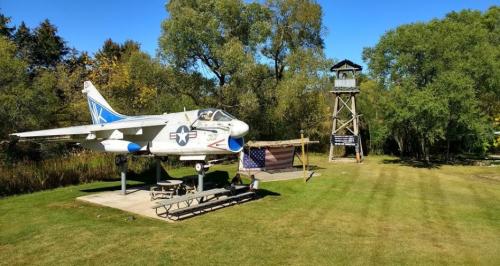 A military jet on display next to a watchtower and picnic area, surrounded by trees and a clear blue sky.