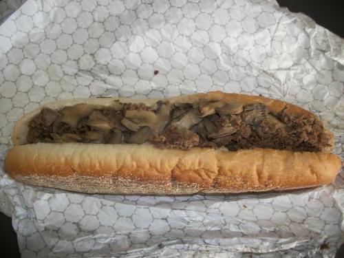 A hoagie roll filled with sliced beef and sautéed mushrooms, wrapped in patterned paper.