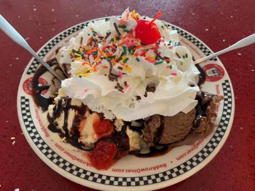 A large ice cream sundae topped with whipped cream, sprinkles, a cherry, and chocolate sauce on a checkered plate.