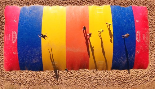Aerial view of people jumping on a colorful inflatable bounce pad on a sandy surface.