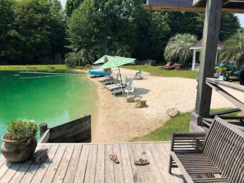 A serene lakeside scene with lounge chairs, a sandy beach, and green trees under a clear sky.