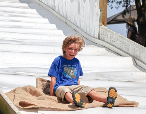 A joyful child with curly hair slides down a large, white slide, laughing as he reaches the bottom.