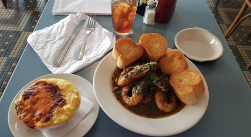 A plate of shrimp in sauce with bread rolls, alongside a dish of cheesy baked casserole and a drink.
