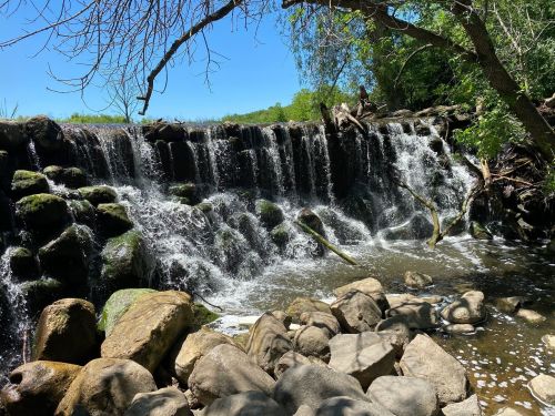 A serene waterfall cascades over rocks, surrounded by lush greenery and a clear blue sky.