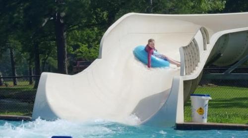 A child slides down a water slide into a pool, splashing water around in a sunny outdoor setting.
