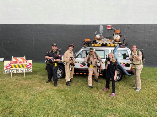 A group of Ghostbusters in costumes poses with a themed vehicle and a sign for Lehigh Valley Ghostbusters.