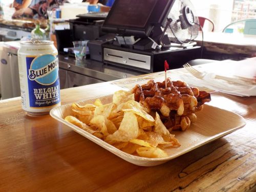 A plate of BBQ chicken topped with a cherry, served with potato chips and a can of Blue Moon beer on a wooden table.