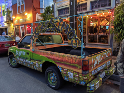 A colorful pickup truck with "Jacques Timos" painted on the side, parked on a lively street with vibrant lights.
