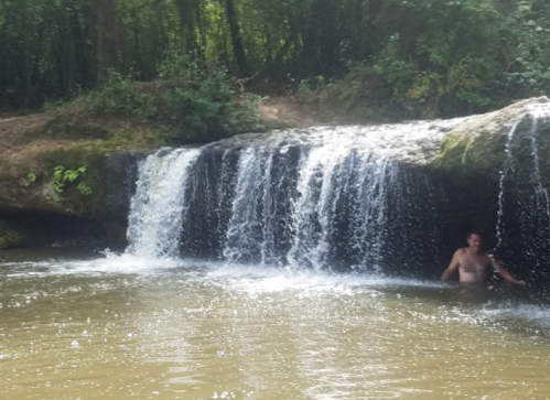 A person stands in a river near a small waterfall, surrounded by lush greenery and rocky terrain.