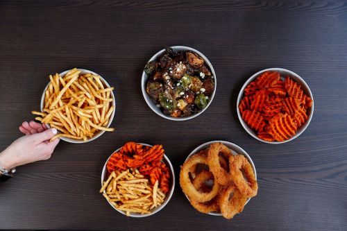A table with five bowls of various fries, onion rings, and roasted vegetables, with a hand reaching for a bowl.