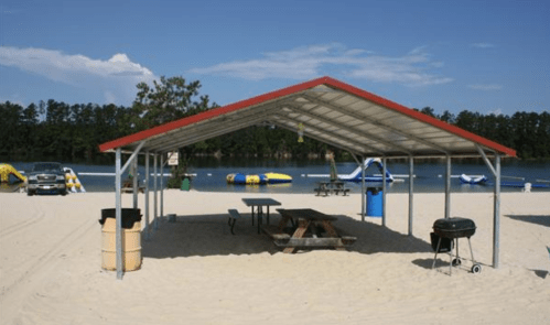 A shaded picnic area with a table and grill on a sandy beach by a lake, featuring inflatable water toys in the background.