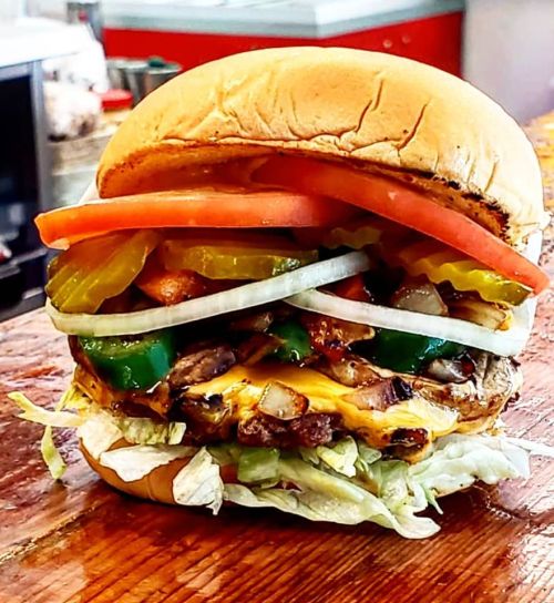 A towering burger with layers of cheese, grilled meat, lettuce, tomatoes, pickles, onions, and green peppers on a bun.