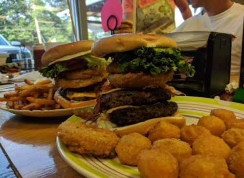 A towering burger with lettuce, bacon, and onion rings, served with fries and tater tots on a colorful plate.