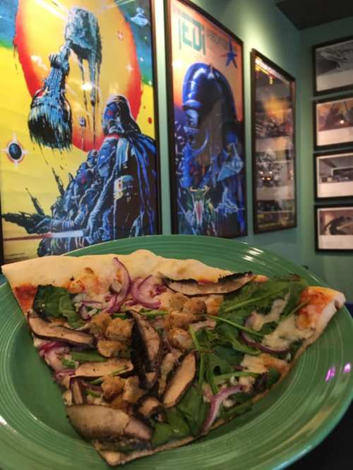 A slice of pizza topped with mushrooms, spinach, and red onions, in front of colorful sci-fi posters on a green wall.