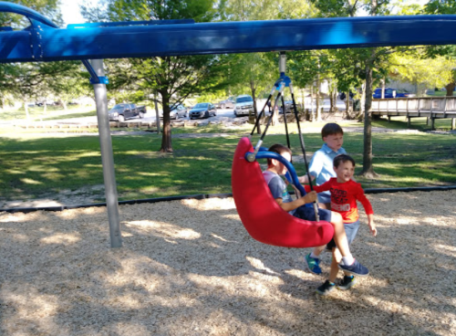 Two children play on a swing at a park, while another child pushes them. Trees and cars are visible in the background.