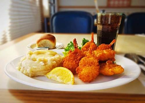 A plate with fried shrimp, mashed potatoes, a lemon wedge, a roll, and a glass of cola on a wooden table.