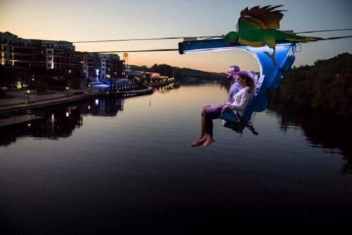 A couple enjoys a scenic zip line ride over a calm river at sunset, with colorful lights illuminating the scene.