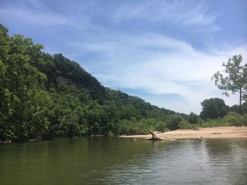 A serene river scene with lush green trees and a rocky hillside under a blue sky with scattered clouds.
